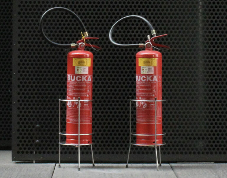 Photo of two fire extinguishers on the floor standing next to each other. The extinguishers are brightly coloured red, labeled with safety instructions and usage guidelines.